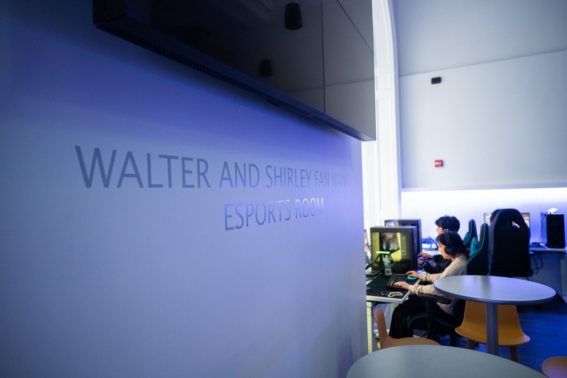 A side shot of the Wallach Esports Lounge, with a TV on the wall about signage, and students sitting at computers playing games.