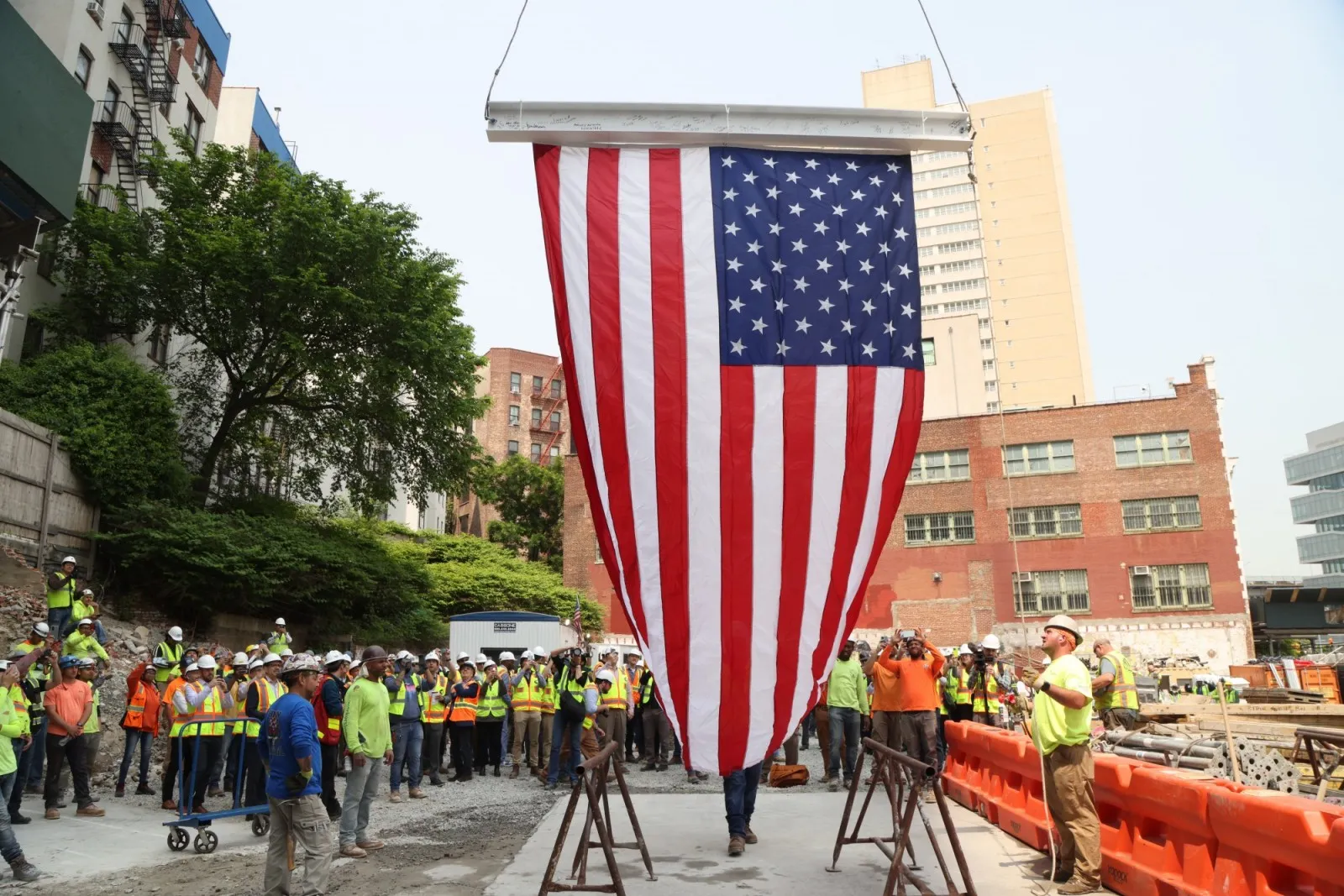 A group of construction workers look on as a signed beam and large flag are hoisted in the air.